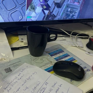 Perfuem - Bilden visar ett skrivbord med en svart kaffemugg, en datormus och papper. En skärm i bakgrunden visar övervakningsbilder.