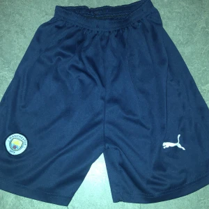 Mörkblå fotbollsshorts från Puma - Snygga mörkblå fotbollsshorts från Puma med Manchester City-logga. Perfekta för fotbollsträningen eller matchen. De har en bekväm passform och är tillverkade i ett lätt och andningsbart material.