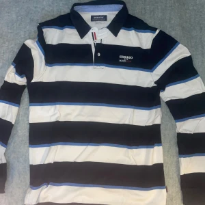 Randig pikétröja från Sebago - Säljer en stilren randig långärmad pikétröja från Sebago i storlek L. Tröjan har breda ränder i svart, vit och blå med broderad logga på bröstet. Den har långa ärmar och en klassisk krage med knappar. Perfekt för en avslappnad och snygg look.