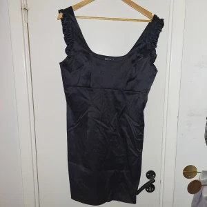 Svart klänning från Gina Tricot - Snygg svart klänning från Gina Tricot med volangdetaljer på axelbanden. Klänningen har en dragkedja på sidan för enkel påklädning. Perfekt för en stilren look.