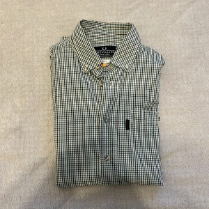 Rutig skjorta från Chevalier - Säljer en klassisk rutig skjorta från Chevalier i grönt och vitt. Skjortan har en normal passform och är perfekt för en stilren look. Den är tillverkad i ett bekvämt material och har knappar framtill.