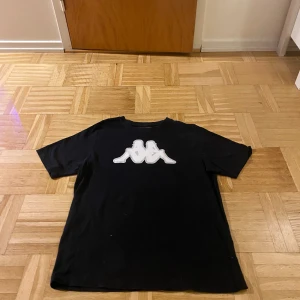 Svart t-shirt från Kappa - Säljer en svart t-shirt från Kappa med det ikoniska logotyptrycket i vitt på bröstet. Perfekt för en avslappnad stil. Tröjan är storlek M