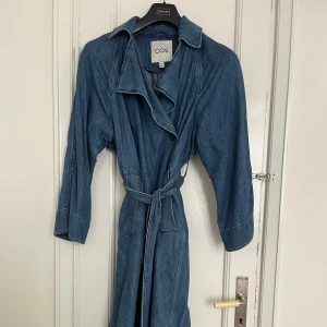 Blå jeans trenchcoat från COS - Snygg blå jeans trenchcoat från COS med knytskärp i midjan. Perfekt för en avslappnad stil med en touch av elegans. Tillverkad i en blandning av bomull och lyocell för en bekväm passform.