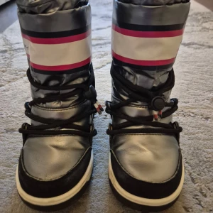 Silvriga moon boots från Tommy Hilfiger - Snygga och varma silvriga moon boots från Tommy Hilfiger med svarta och vita detaljer. De har en cool design med rosa och svarta ränder upptill och snörning framtill. Perfekta för vinterns kalla dagar! 🥶