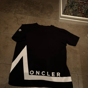 Svart t-shirt från Moncler - Snygg svart t-shirt från Moncler med vit logga och grafisk design på framsidan. T-shirten har korta ärmar och en klassisk rund halsringning. Perfekt för en stilren look.