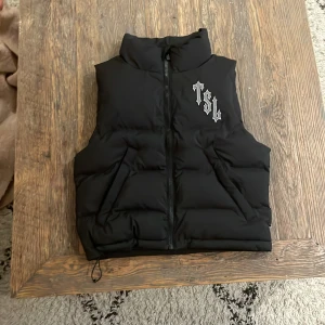 Svart dunväst från Trapstar - Säljer en svart dunväst från Trapstar med dragkedja och broderad logga på bröstet. Baksidan har ett stort tryck med texten 'Trapstar London Shooters'. Västen är tillverkad i 100% polyester och passar perfekt för kyligare dagar.