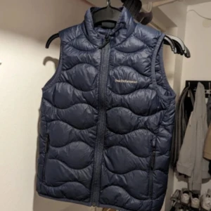 Blå dunväst från Peak Performance, strl 150 - Säljer en snygg blå dunväst från Peak Performance i jättefint skick. Västen har en dragkedja framtill och är quiltad för extra värme. Ser helt ny ut. 