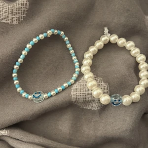 Två armband med pärlor och smiley - Säljer två armband, ett med vita och blå pärlor och ett med större vita pärlor. Båda har en charmig smiley-detalj i blått och vitt. Perfekta för att ge en lekfull touch till din stil!