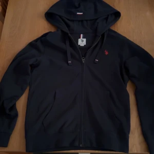 Mörk blå från U.S. Polo Assn. - Säljer en stilren mörkblå hoodie från U.S. Polo Assn. med dragkedja och röd broderad logga på bröstet. Perfekt för en avslappnad look med justerbar huva och bekväma muddar. Skriv gärna om ni har frågor och funderingar.