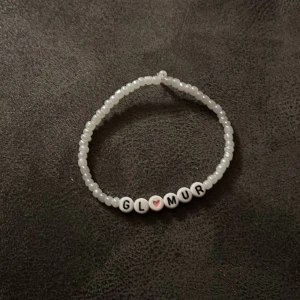 Vitt pärlarmband med text - Charmigt vitt pärlarmband med bokstavspärlor som bildar ordet 'GLAMUR'. En söt rosa hjärtpärla ger en extra touch. Perfekt för att ge en personlig stil till din outfit.