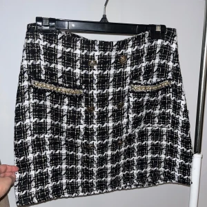 Rutig kjol med knappar - Snygg rutig kjol i svart och vit med dekorativa knappar framtill. Kjolen har en klassisk stil med två fickor och en elegant passform. Perfekt för att ge en chic touch till din outfit.