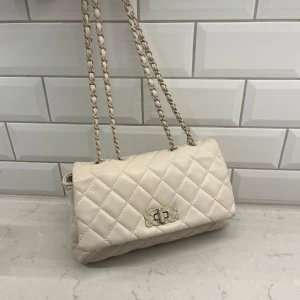 Beige quiltad axelväska - Snygg beige axelväska med quiltat mönster och kedjedetaljer. Väskan har en elegant vridlåsdetalj framtill och en praktisk storlek för vardagligt bruk. Köpt i Spanien för 35€, cirka 400 kronor. Använd så säljer för billigt pris.