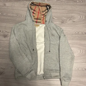 Grå huvtröja Burberry - Säljer en stilren grå huvtröja från Burberry med klassiskt rutigt mönster i fodret. Tröjan har dragkedja och justerbar huva med snören. Perfekt för en avslappnad och trendig look.