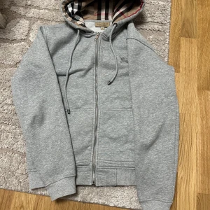 Grå hoodie från Burberry - Säljer en snygg grå hoodie från Burberry i mycket bra skick. Den har en klassisk dragkedja och en huva med det ikoniska Burberry-mönstret på insidan. Perfekt för höst och vår, och den är supermjuk och bekväm. Passar både till vardags och lite finare tillfällen. 🧥✨