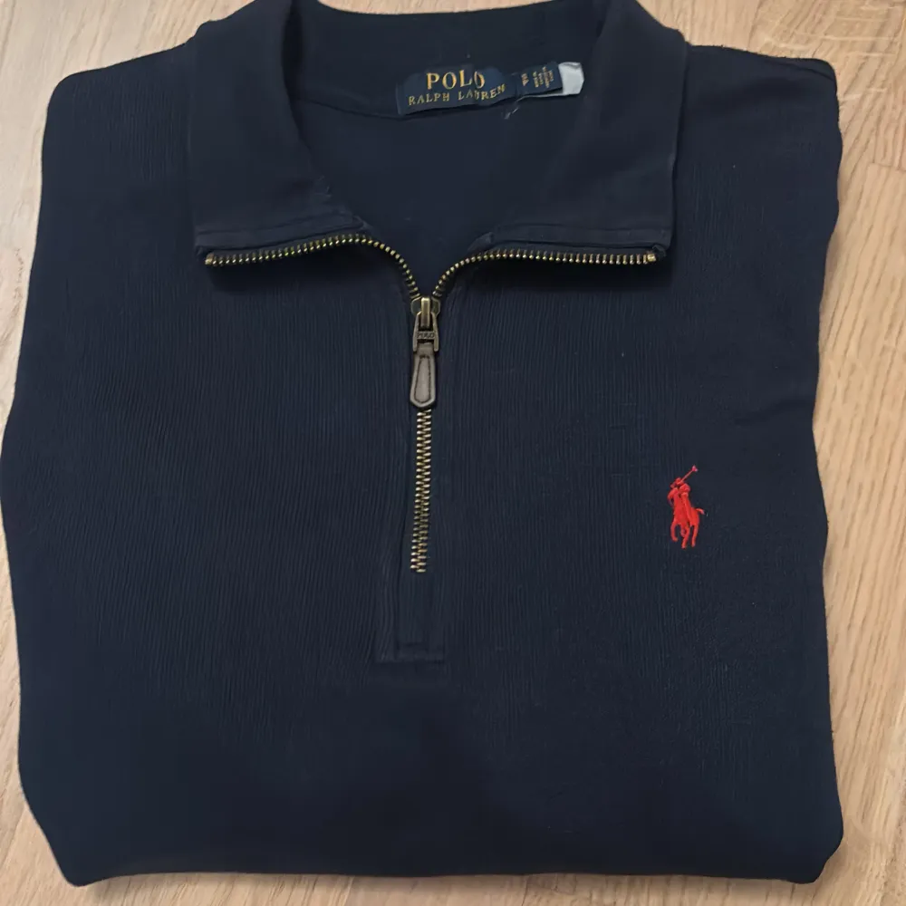 Säljer en stilren navy blue långärmad tröja från Ralph Lauren med en klassisk röd logga på bröstet. Tröjan har en praktisk dragkedja vid halsen och är perfekt för både vardag och lite finare tillfällen. Passar bra till höst och vinter. Skön och bekväm att ha på sig!. Tröjor & Koftor.