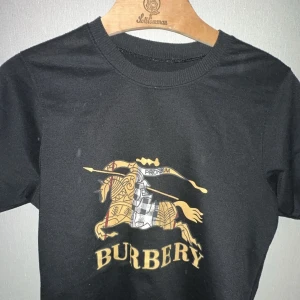 Svart t-shirt från Burberry - Säljer en snygg svart t-shirt från Burberry med ett coolt tryck av en riddare på häst i guld och silver. Perfekt för att ge din outfit en lyxig touch. T-shirten är kortärmad och i bomull, vilket gör den både bekväm och stilren. Passar perfekt till både vardag och fest! 