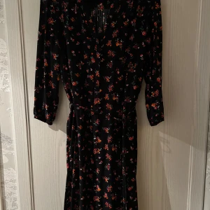 Blommig klänning från Saint Tropez - Säljer en superfin svart klänning från Saint Tropez med färgglatt blommönster. Den har trekvartsärmar och ett knytband i midjan för en smickrande passform. Perfekt för både vardag och fest!