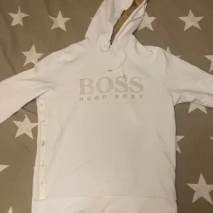  Hugo Boss hoody - Säljer en stilren vit hoodie från Hugo Boss. Den har en klassisk design med logotypen tryckt på framsidan och en bekväm passform. Perfekt för både vardag och avslappnade tillfällen. Huvan har dragsko och tröjan är i mycket bra skick.