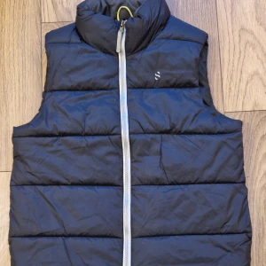 Svart dunväst från H&M Sport - Säljer en svart dunväst från H&M Sport med dragkedja framtill. Västen är quiltad och har en hög krage för extra värme. Perfekt för kyliga dagar.