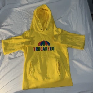 Gul hoodie från Trocadero - Säljer en cool gul hoodie från Trocadero med färgglatt tryck på framsidan. Hoodien har en klassisk design med känguruficka. Perfekt för en avslappnad stil!