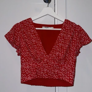 Röd blommig croppad topp från NA-KD - Säljer en söt röd croppad topp från NA-KD med vita blommor. Toppen har en v-ringad design och korta ärmar samt resor i midjan baktill på tröjan.