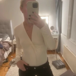 Vit långärmad body  - Säljer en stilren vit långärmad body med v-ringning och en snygg knytdetalj framtill. Perfekt för både vardag och festliga tillfällen. Toppen har en tight passform som framhäver figuren. Passar bra till både jeans och kjol!