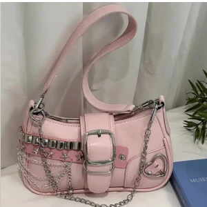 Rosa axelväska med kedjedetaljer - Snygg rosa axelväska i syntetmaterial med coola kedjedetaljer och stjärnprydda nitar. Väskan har ett hjärtdetalj och ett bälte med spänne framtill. Perfekt för att ge din outfit en edgy touch.