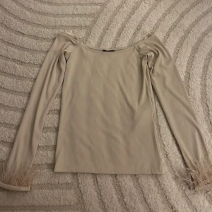 Beige topp med fjäderdetaljer - Säljer en stilren beige topp med långa ärmar och unika fjäderdetaljer vid ärmsluten. Perfekt för att ge en touch av elegans till din outfit. Toppen har en mjuk och bekväm passform.