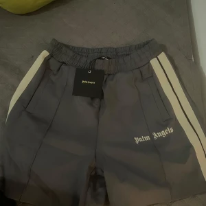 Grå shorts från Palm Angels - Säljer ett par grå shorts från Palm Angels i nytt skick. De har en bekväm elastisk midja och vita sidoremsor som ger en sportig look. Perfekta för både träning och avslappnade dagar. Märkesloggan är snyggt placerad på ena benet. Köpt är köpt lämna ej tbx 