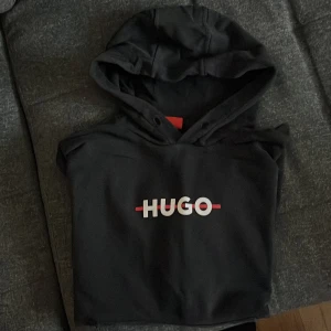 Svart hoodie från Hugo - Säljer en stilren svart hoodie från Hugo med logga i vitt och rött på bröstet. Tröjan har en klassisk passform med långa armar och en praktisk magficka. Perfekt för en avslappnad look. Den är i bra skick, pris kan diskuteras vid snabbafä. 