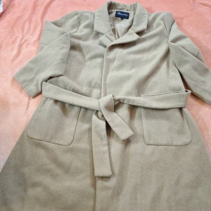 Beige kappa från Happy Holly - Elegant beige kappa från Happy Holly med knytbälte i midjan. Den har en klassisk design med långa ärmar och knappar framtill. Perfekt för höst och vår.