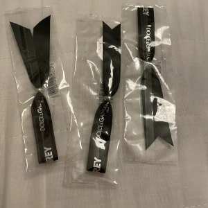 Svart parfymband från Dolce & Gabbana - Svart satinband från Dolce & Gabbana med texten 'The One' tryckt i vitt. Perfekt för att doftsätta med din favoritparfym. Bandet är elegant och stilrent, idealiskt för att ge en lyxig touch till presentinslagningar eller som accessoar.
