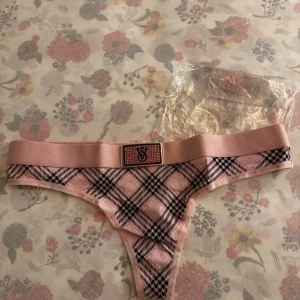 Rutig rosa trosa från Victoria's Secret - Snygg rosa trosa från Victoria's Secret med ett rutigt mönster i svart och rosa. Trosan har en bred resår med en glittrig detalj framtill. Perfekt för att känna sig både bekväm och stilren.