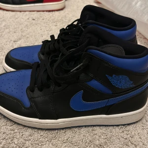Jordans 1 mid hyper royal - Snygga Nike Air Jordans i svart och blått med klassisk snörning. Skorna har en hög design med den ikoniska Air Jordan-loggan på sidan och tungan. Perfekta för en sportig look. 