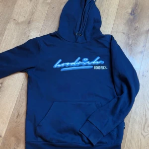 Blå hoodie från Hoodrich - Säljer en blå hoodie från Hoodrich med blått och vitt tryck på framsidan. Den har en klassisk känguruficka och justerbar huva med dragsko. Perfekt för streetwear-stilen. Finns även en gömd ficka i ena ärmen. Tröjan är i storlek S och passar dig som är 170-180