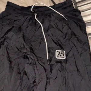 Svarta vind byxor från EA7 Emporio Armani S/M - Säljer ett par svarta Windbreakers från EA7 Emporio Armani. De har en bekväm passform med dragsko i midjan för justerbarhet. Perfekta för både träning och avslappnade dagar. Märkeslogga på benet ger en stilren touch. Äkta med qr kod som tar en till hemsidan