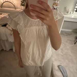 Vit blus med spetsdetaljer - Säljer en söt vit blus med spetsdetaljer och volangärmar. Perfekt för sommardagar eller en avslappnad vårlook. Blusen har en luftig passform och är i mycket bra skick. Passar både till jeans och kjol för en chic stil.