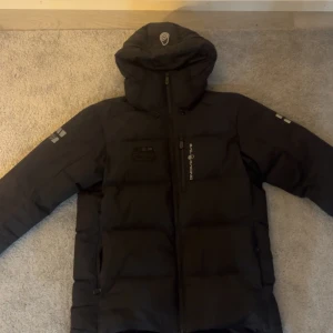 Svart dunjacka från Sail Racing - Säljer en svart dunjacka från Sail Racing i mycket bra skick. Jackan är vindtät och andas bra, perfekt för kalla vinterdagar. Den har en dragkedja framtill och är långärmad. Jackan är fylld med RDS-certifierat dun och har en DWR-behandling för extra skydd mot väta. Perfekt för dig som vill hålla dig varm och stilren i vinter! Har bara haft den en vinter och den är helt ny!