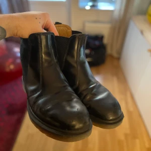 Svarta Chelsea boots från Dr. Martens - Säljer ett par klassiska svarta Chelsea boots från Dr. Martens. Skorna är tillverkade i skinn och har en elastisk panel på sidan för enkel på- och avtagning. Perfekta för både vardag och mer uppklädda tillfällen.