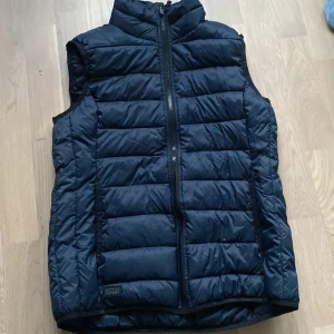 Svart quiltad dunväst från South West - Säljer en svart quiltad dunväst från South West med dragkedja framtill. Perfekt för kyligare dagar och enkel att matcha med andra plagg. Västen har en stilren design och är i storlek medium.