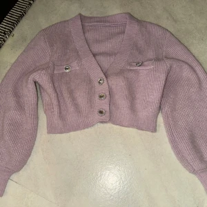 Lila stickad kofta med glittriga knappar - Säljer en söt lila stickad kofta med glittriga knappar framtill. Koftan är croppad och har långa ärmar med ribbade muddar. Perfekt för att lägga till lite glitter i vardagen!