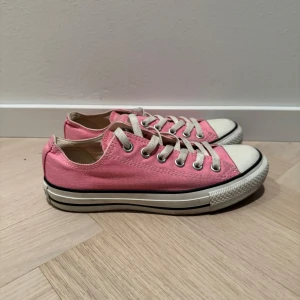 Rosa sneakers från Converse - Säljer ett par klassiska rosa Converse All Star sneakers med vita snören och tåhätta. Skorna har en låg profil och den ikoniska loggan på hälen. Perfekta för en avslappnad stil.