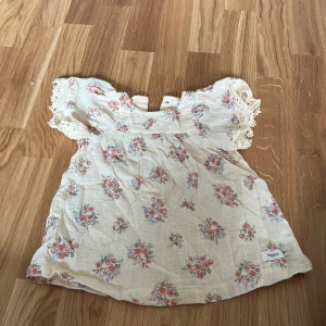 Blommig klänning från Newbie - Söt blommig klänning från Newbie i storlek 68 för 4-6 månader. Klänningen har korta ärmar med spetsdetaljer och ett vackert blommönster i rosa och blått på en beige bakgrund. Perfekt för sommarens varma dagar.