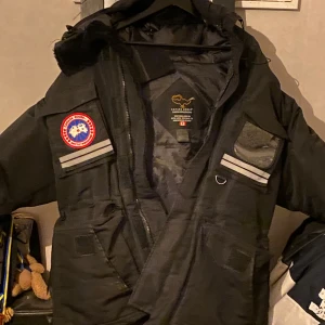 Svart parkajacka från Canada Goose - Säljer en svart parkajacka från Canada Goose, modell Snow Mantra. Jackan har en huva och flera praktiska fickor. Den är dekorerad med en tydlig logotyp på ärmen och har reflexdetaljer för extra synlighet. Perfekt för kalla vinterdagar.  dragkedjan har försvunnit därför säljer jag den billigare. Pris går att diskutera såklart!