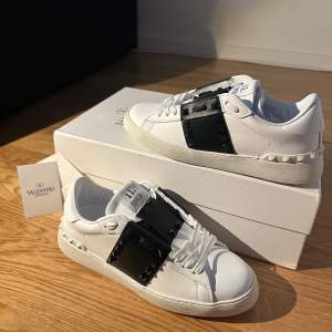 Snygga Valentino Open sneakers i vitt skinn med svarta detaljer. Skorna har en unik design med svarta nitar och snörning. Perfekta för en stilren look.