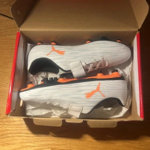 Vita fotbollsskor från Puma - Säljer ett par snygga vita fotbollsskor från Puma i nytt skick. De har en cool orange detalj på sidan och svarta sulor med orange dobbar. Skorna har både snörning och kardborreband för bästa passform. Perfekta för fotbollsträning eller match!