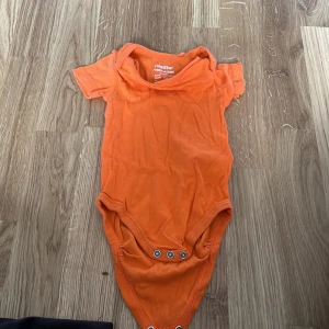 Orange kortärmad body från Lupilu - Säljer en söt orange kortärmad body från Lupilu. Perfekt för små barn, med praktiska knappar i grenen för enkel på- och avklädning. Mjuk och bekväm för lek och vila.