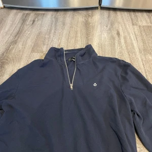 Mörkblå tröja med dragkedja - Säljer en stilren mörkblå tröja med dragkedja och liten logga på bröstet. Perfekt för kyligare dagar och passar till det mesta. Tröjan har långa ärmar och en hög krage.