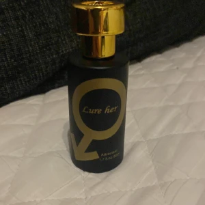 Lure Her Attracion Parfym - Elegant parfymflaska i svart med gyllene detaljer och texten 'Lure her'. Flaskan har en lyxig gyllene kork och rymmer 50 ml av doften Attracion.