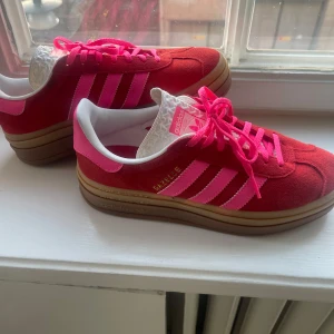Röda Adidas Gazelle sneakers - Säljer ett par snygga röda Adidas Gazelle sneakers i mocka med rosa detaljer och snörning. De har en klassisk design med gummisula och är i nytt skick. Perfekta för både vardag och fest! 🏃‍♂️✨